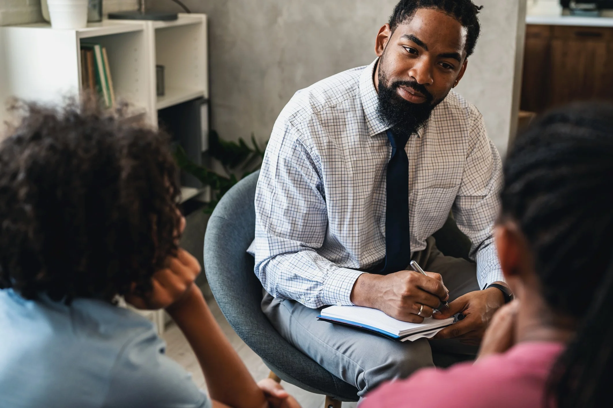 Man in checkered shirt and tie sitting, surrounded by women, engaged in discussion during meeting or group conversation.