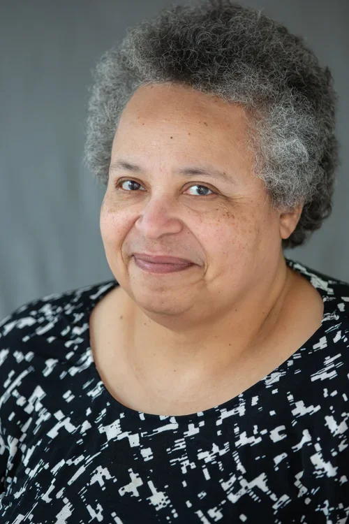 A close-up portrait of an older woman with short, curly gray hair, smiling gently, wearing a black and white patterned top.