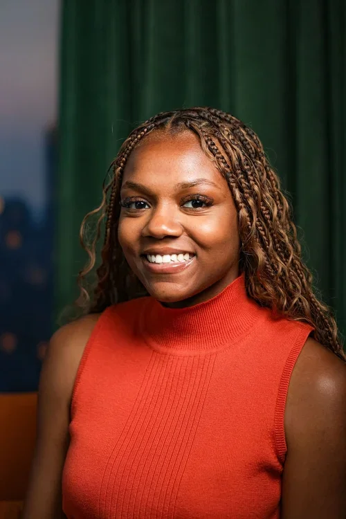 Portrait of a smiling woman with braided hair wearing an orange sleeveless top, standing in front of a green curtain.