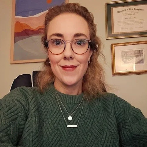 A woman with glasses, curly hair, and a green sweater, smiling for a selfie in a room with framed diplomas on the wall.
