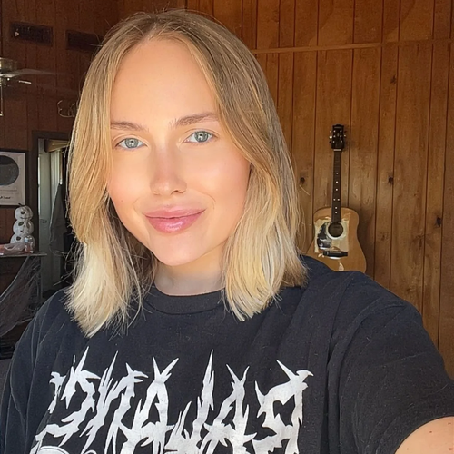 A young woman with blond hair and blue eyes taking a selfie in a room with wooden walls. She is wearing a black T-shirt with white graphic text and has a slight smile. In the background, there is a guitar hanging on the wall.