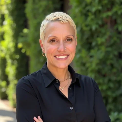 A woman with short blonde hair smiling outdoors in front of green foliage, wearing a black collared shirt and gold jewelry.