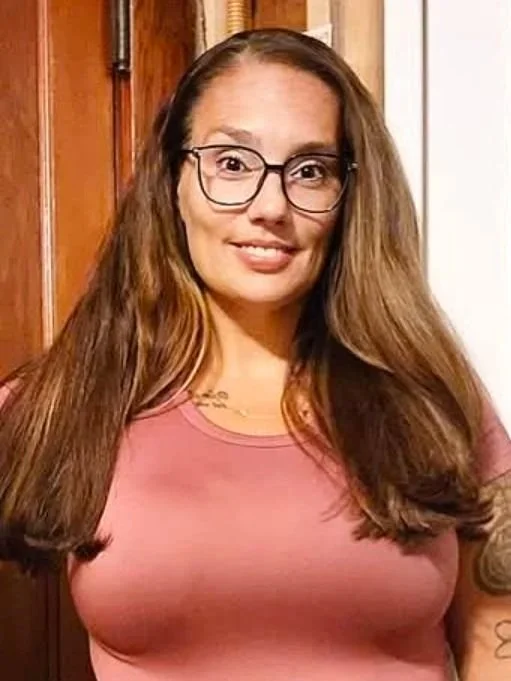 A woman with long brown hair, wearing glasses and a pink shirt, smiling at the camera in a room with wooden doors.