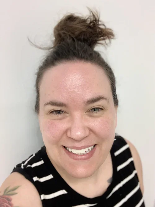 A woman with light skin, brown hair tied in a messy bun, wearing a black and white striped sleeveless top, smiling and looking at the camera.