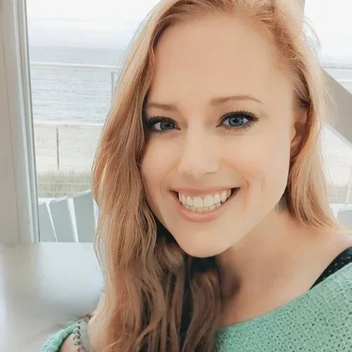 A young woman with long red hair and blue eyes smiling in front of a beach view.