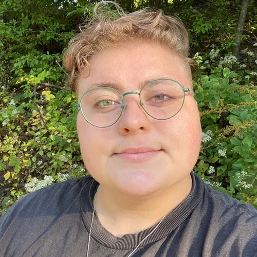 Katie Little of Aviv is tra Domestic & Intimate Partner Violence, Trauma, Substance Use Disorders/ Harm Reduction, LGBTQ+ youth experience with short curly hair wearing glasses.