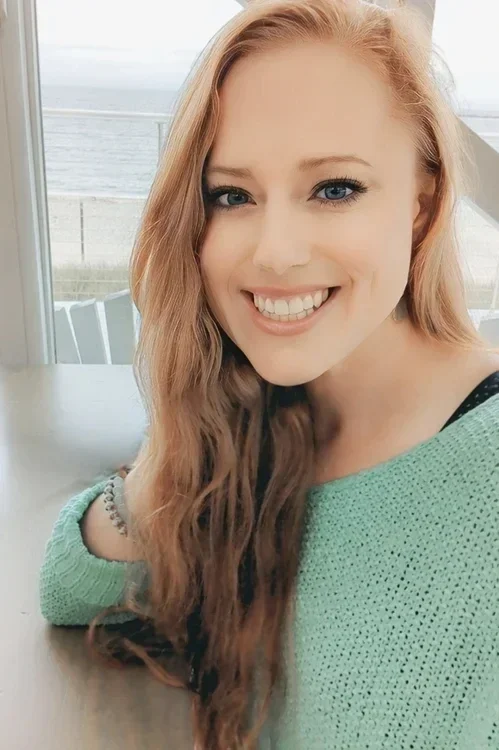 A woman with long, wavy red hair and blue eyes smiling while sitting indoors near a window with a view of the ocean in the background.