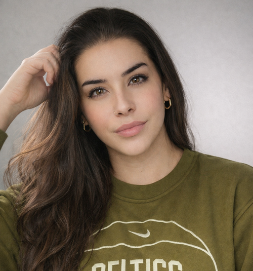 Young woman with long brown hair, wearing a green sweatshirt and gold hoop earrings, posing in front of a neutral background
