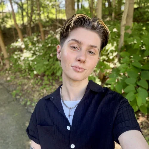 Young person with short hair and earrings taking a selfie outdoors with green trees and bushes in the background.