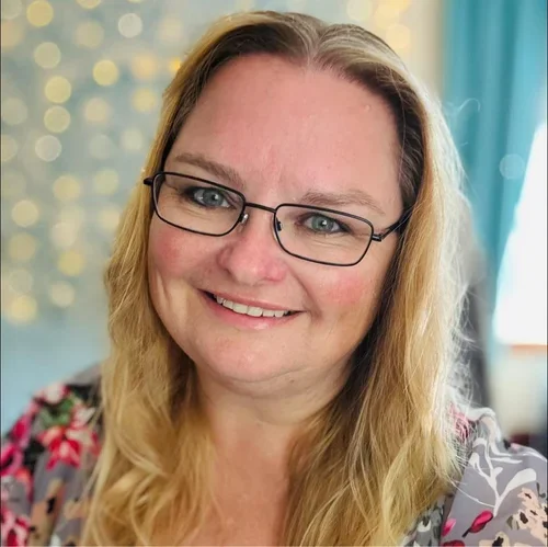 A smiling woman with glasses and long blonde hair, indoors with a blurred background of colorful lights and curtains.
