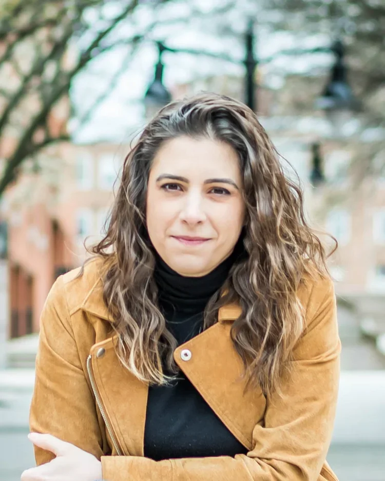 A woman with curly brown hair, wearing a black turtleneck and tan jacket, stands outdoors with trees and buildings in the background.