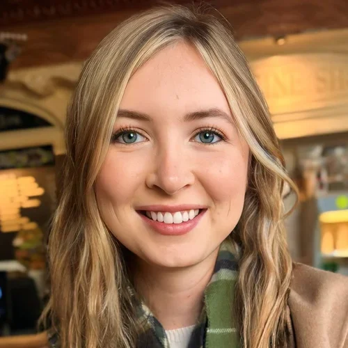 A smiling young woman with long blonde hair and blue eyes in a cozy indoor setting.