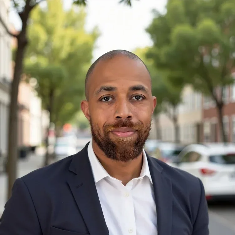 A man with a shaved head and beard wearing a dark suit jacket and white shirt, standing outdoors on a city street with trees and parked cars in the background.