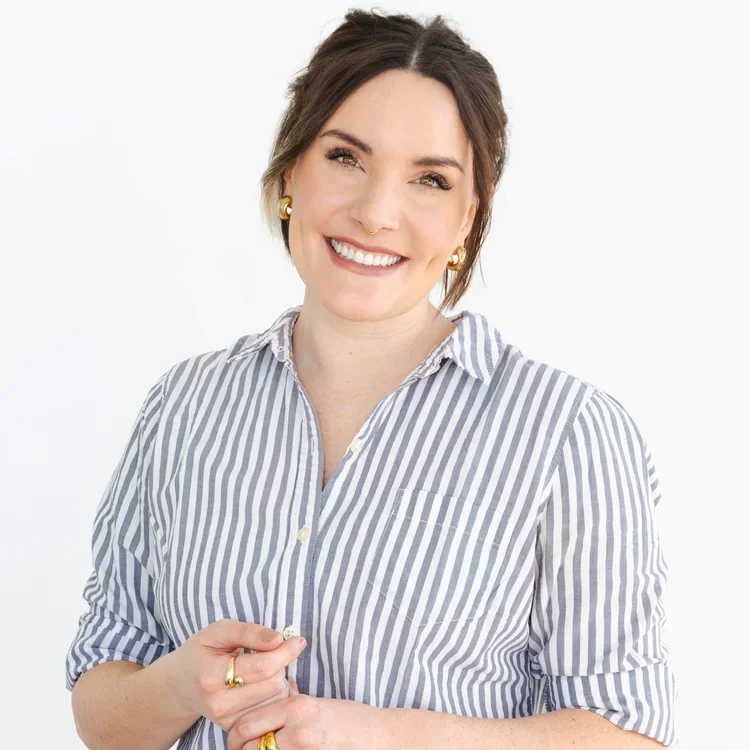 A woman with short brown hair, wearing a striped button-down shirt, smiling, with gold earrings and rings, standing against a white background.