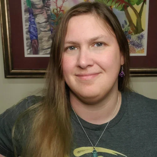 Portrait of a woman with long brown hair wearing a dark gray shirt, standing in front of colorful framed artwork on the wall.
