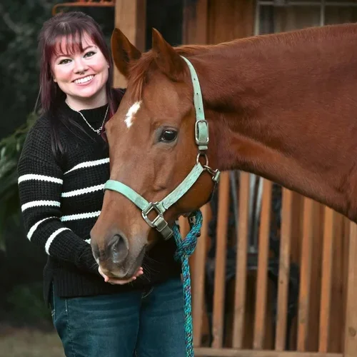 A woman with red hair in a black sweater standing next to a brown horse with a turquoise halter and lead rope, outdoors near a wooden structure.