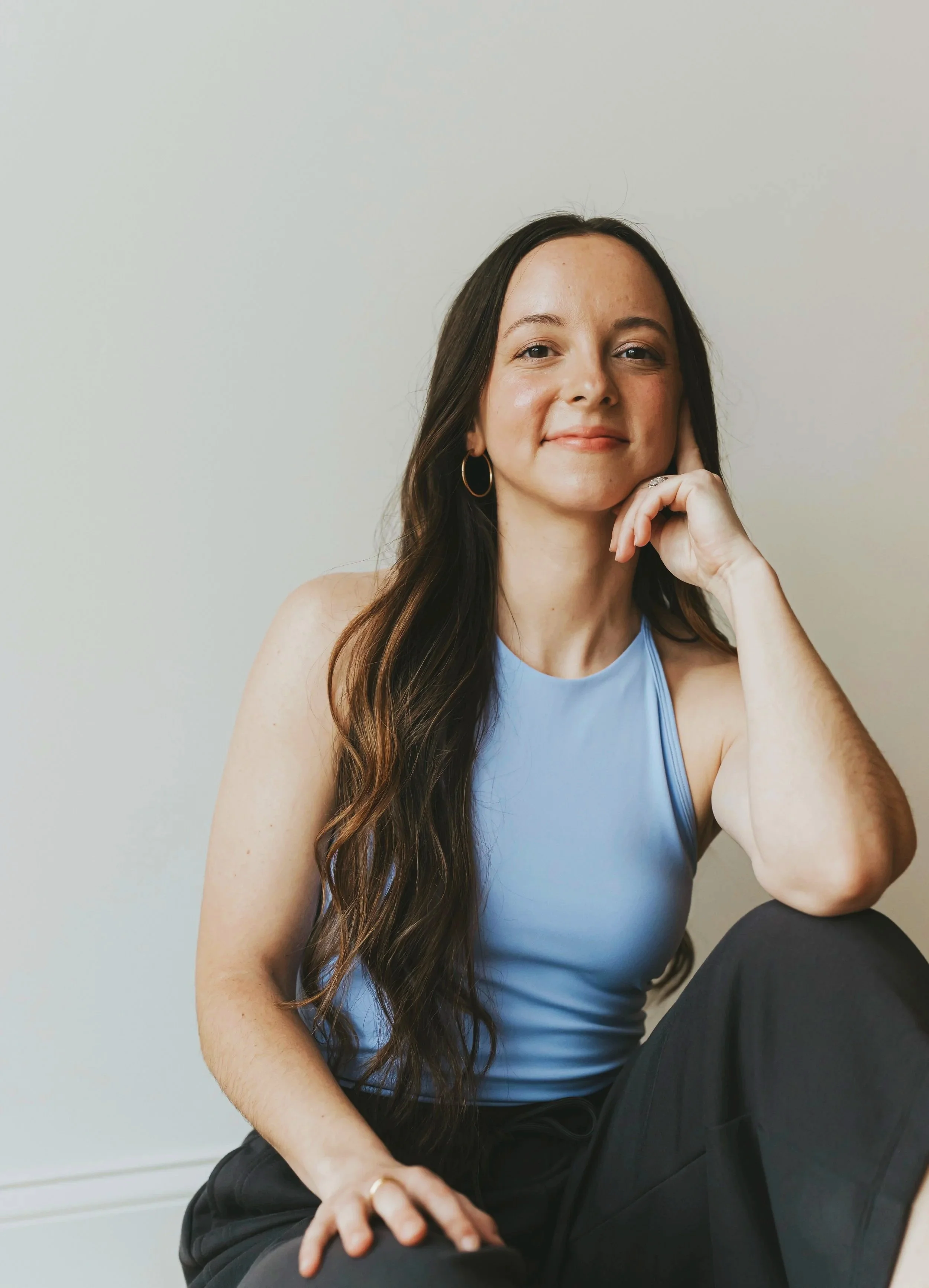 A woman with long brown hair and hoop earrings sitting against a plain light-colored wall, smiling gently with her head resting on her hand.