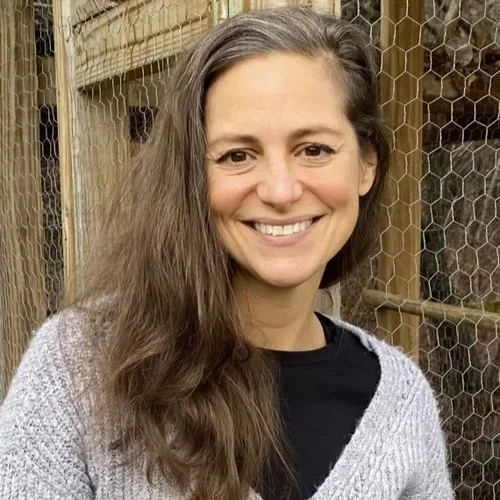 A woman with long brown hair smiling outdoors in front of a chicken wire fence and wooden structure.