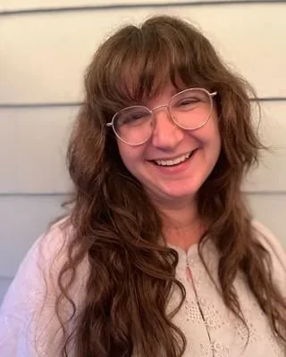 Young woman with long, wavy brown hair and glasses smiling