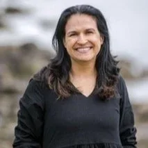 A woman with long dark hair smiling outdoors against a blurred natural background.