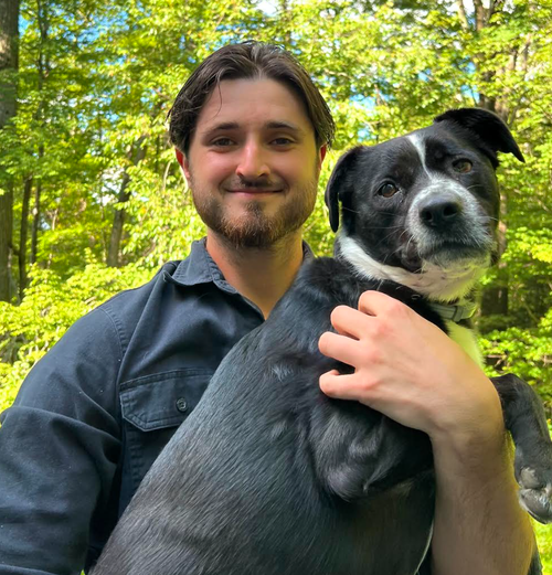 A man with dark hair and a beard smiling while holding a black and white dog outdoors in a wooded area.
