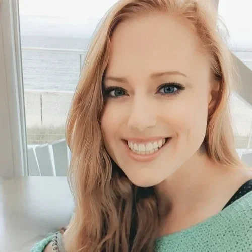 A woman with long blonde hair and blue eyes smiling, sitting indoors near a window with an ocean view.