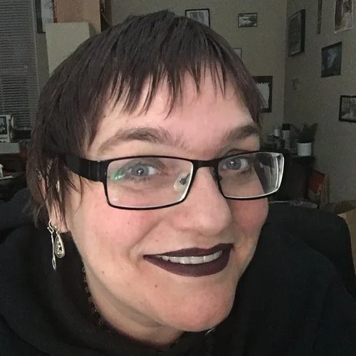 A woman with short brown hair, glasses, and dark lipstick smiling at the camera indoors.