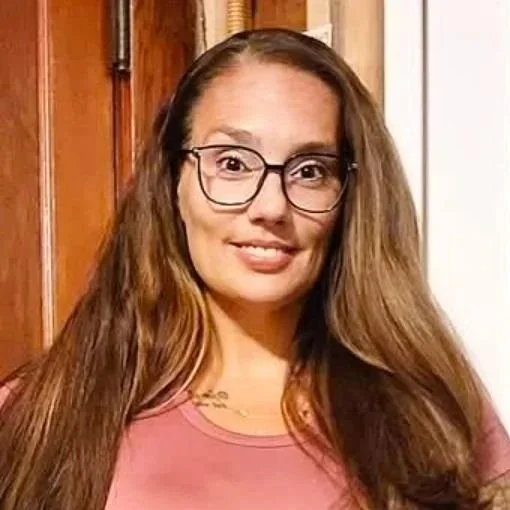 A woman with long brown hair, glasses, and a pink shirt smiling indoors with wooden paneling and a white wall in the background.