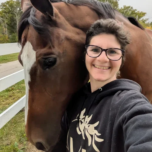 Person taking a selfie with a horse outdoors near a white fence, smiling and wearing glasses and a hoodie.