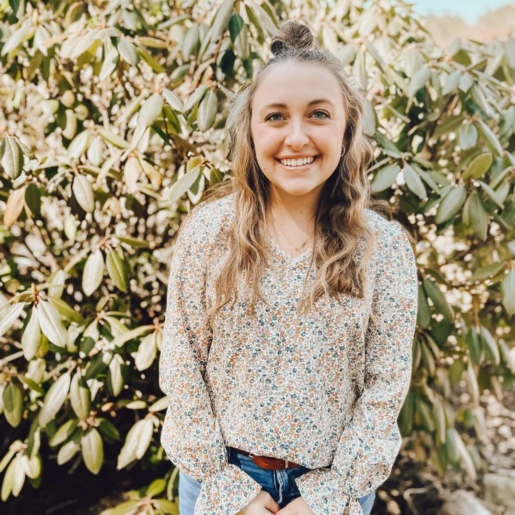 A young woman with long, curly hair tied in a half-up bun, smiling, standing outdoors in front of leafy bushes.