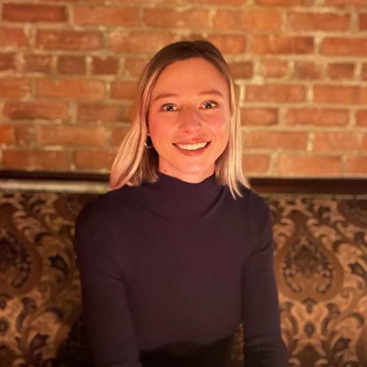 A smiling woman with shoulder-length blonde hair, wearing a black turtleneck sweater, sitting against a brick wall background.
