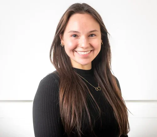 Young woman with long brown hair smiling, wearing a black top and gold jewelry, against a plain white background.