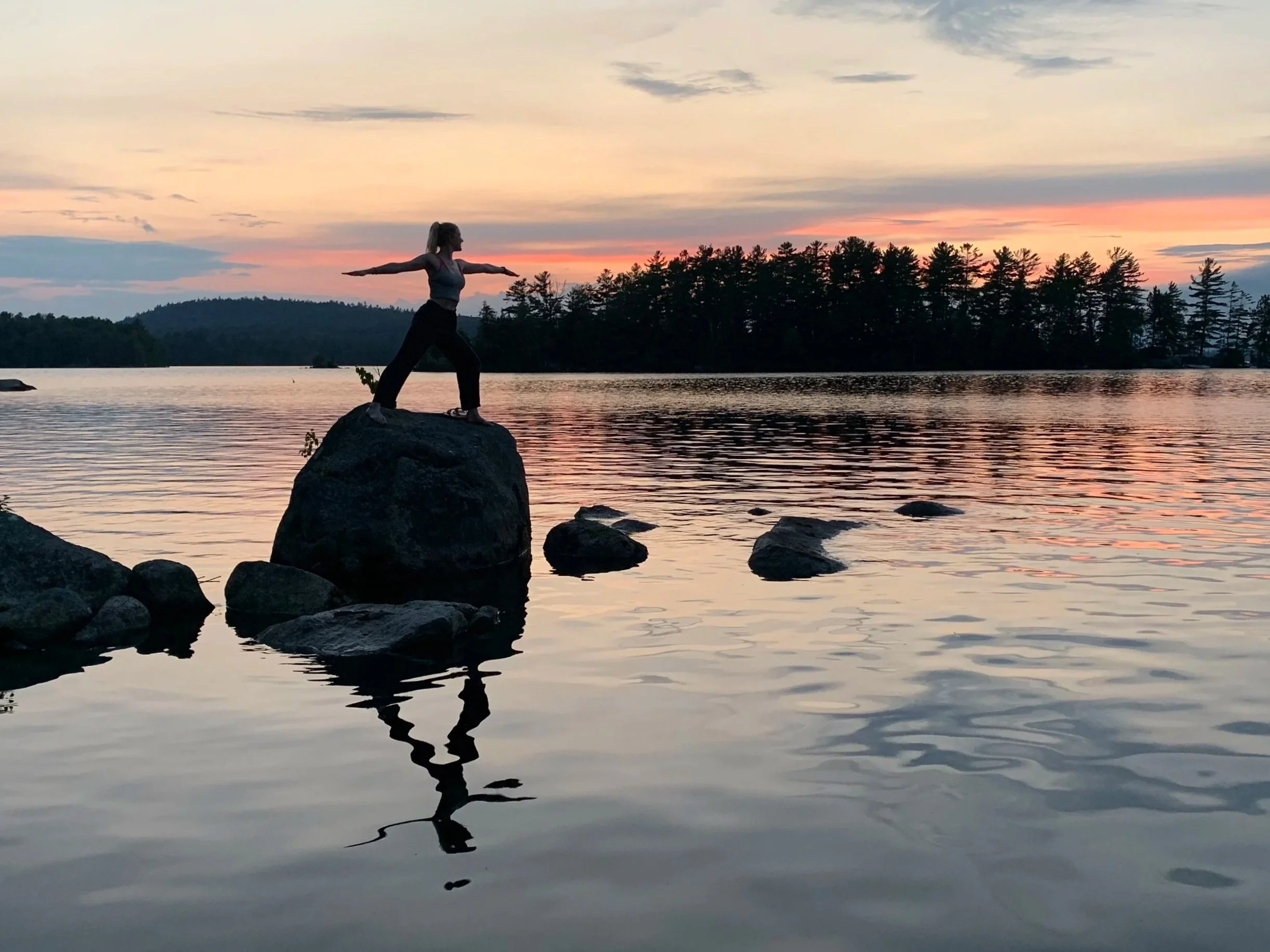 A person practicing yoga on a large rock in a serene lake during sunset, with calm water reflecting the sky and dark silhouette of trees in the background.
