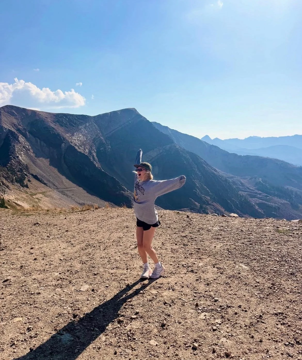 A woman in a gray sweatshirt, black shorts, and white shoes, standing outdoors on a rough terrain, posing with arms raised and a big smile, with mountain ranges and a blue sky in the background.