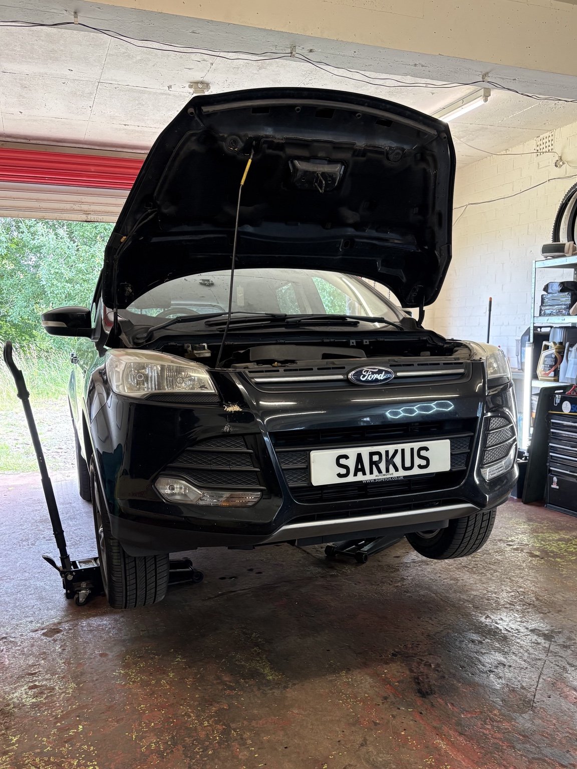 Black Ford SUV with open hood, parked inside a garage, front wheels on a hydraulic lift, with a custom license plate reading 'SARKUS'.