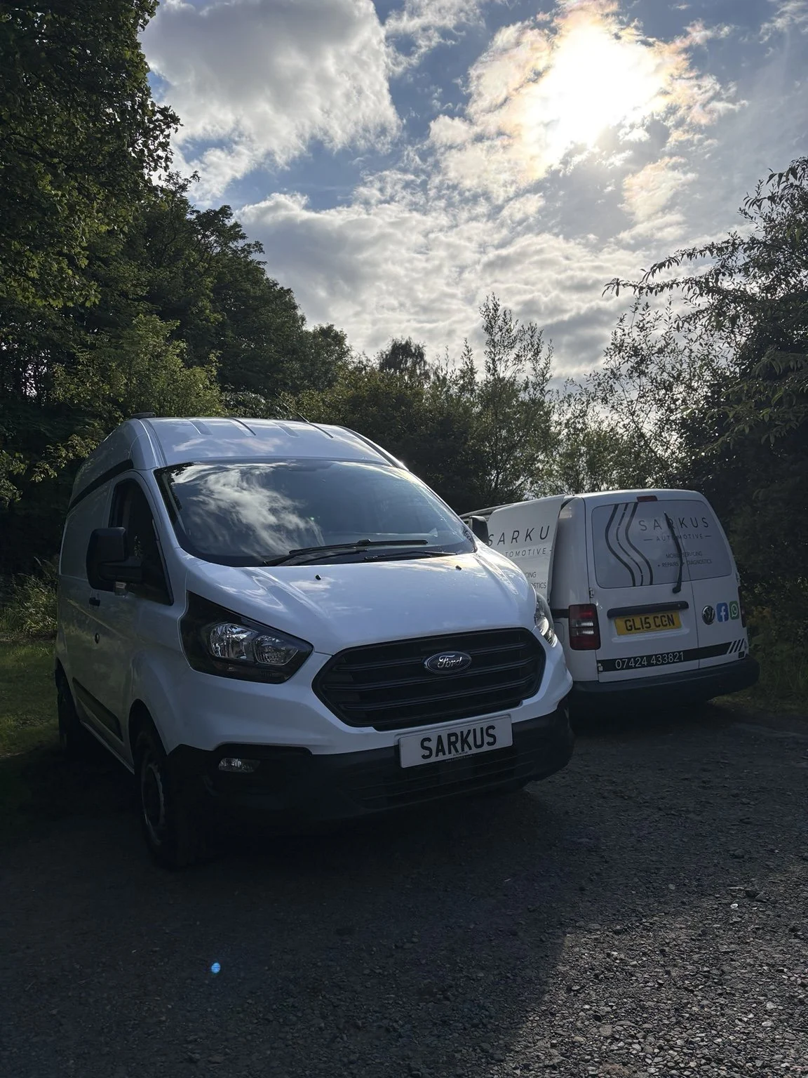 Two white commercial vans parked on a gravel surface with trees and a partly cloudy sky with the sun in the background.