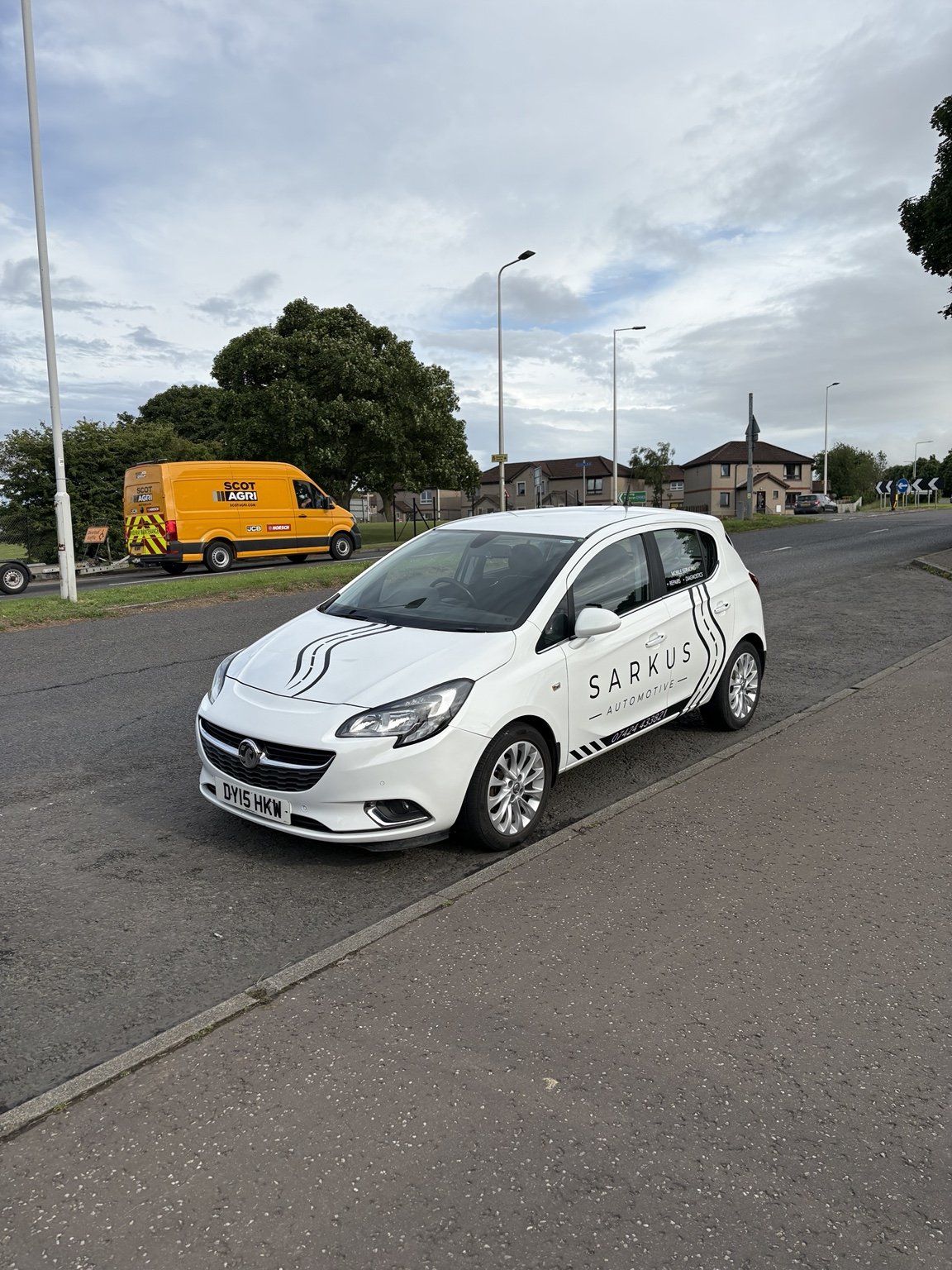 White hatchback car with black racing stripes and 'SARKUS AUTOMOTIVE' branding parked on the side of a road, with an orange utility van and a few houses in the background under a cloudy sky.