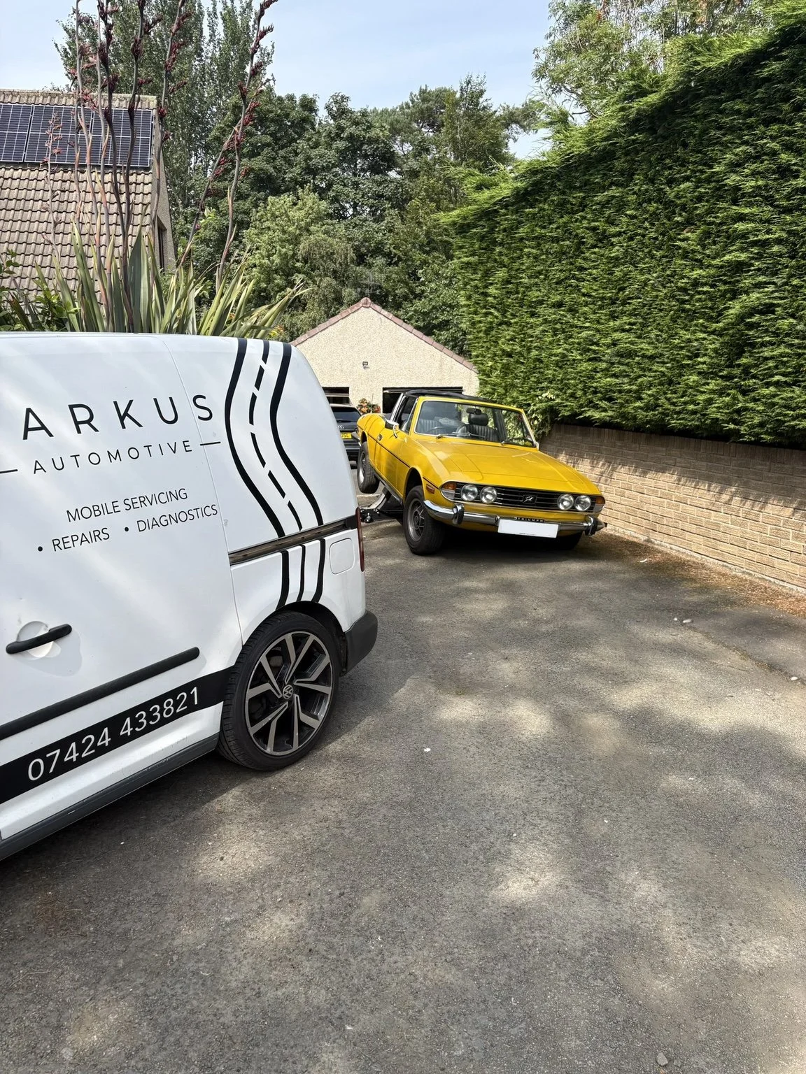 A yellow vintage convertible car parked on a driveway next to a white vehicle with automotive services branding. The driveway is bordered by a green hedge and a brick wall, with trees and a house in the background.