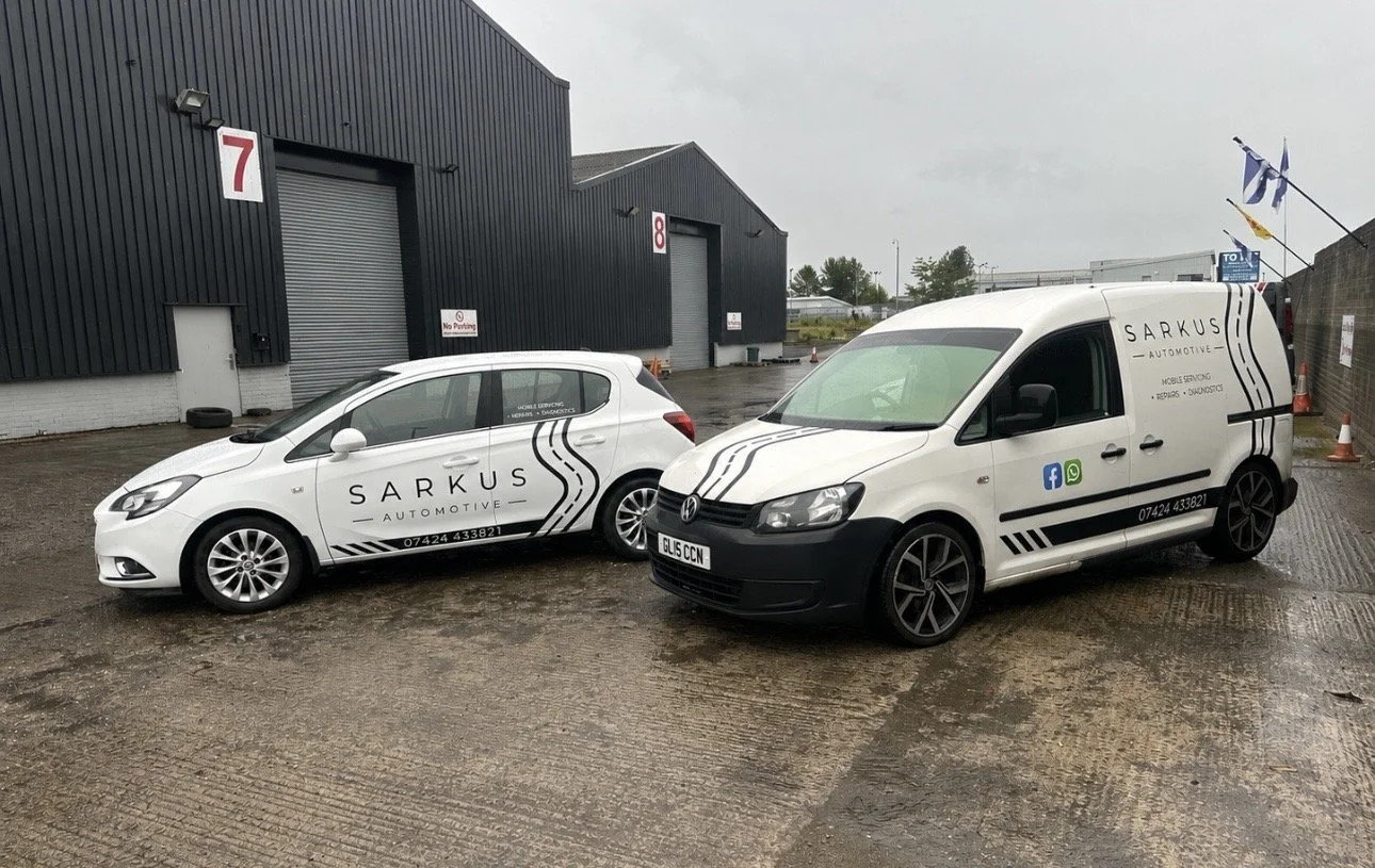 Two white service vehicles branded with Sarkus Automotive logo parked outside a building with two garage doors numbered 7 and 8, on a wet and overcast day.
