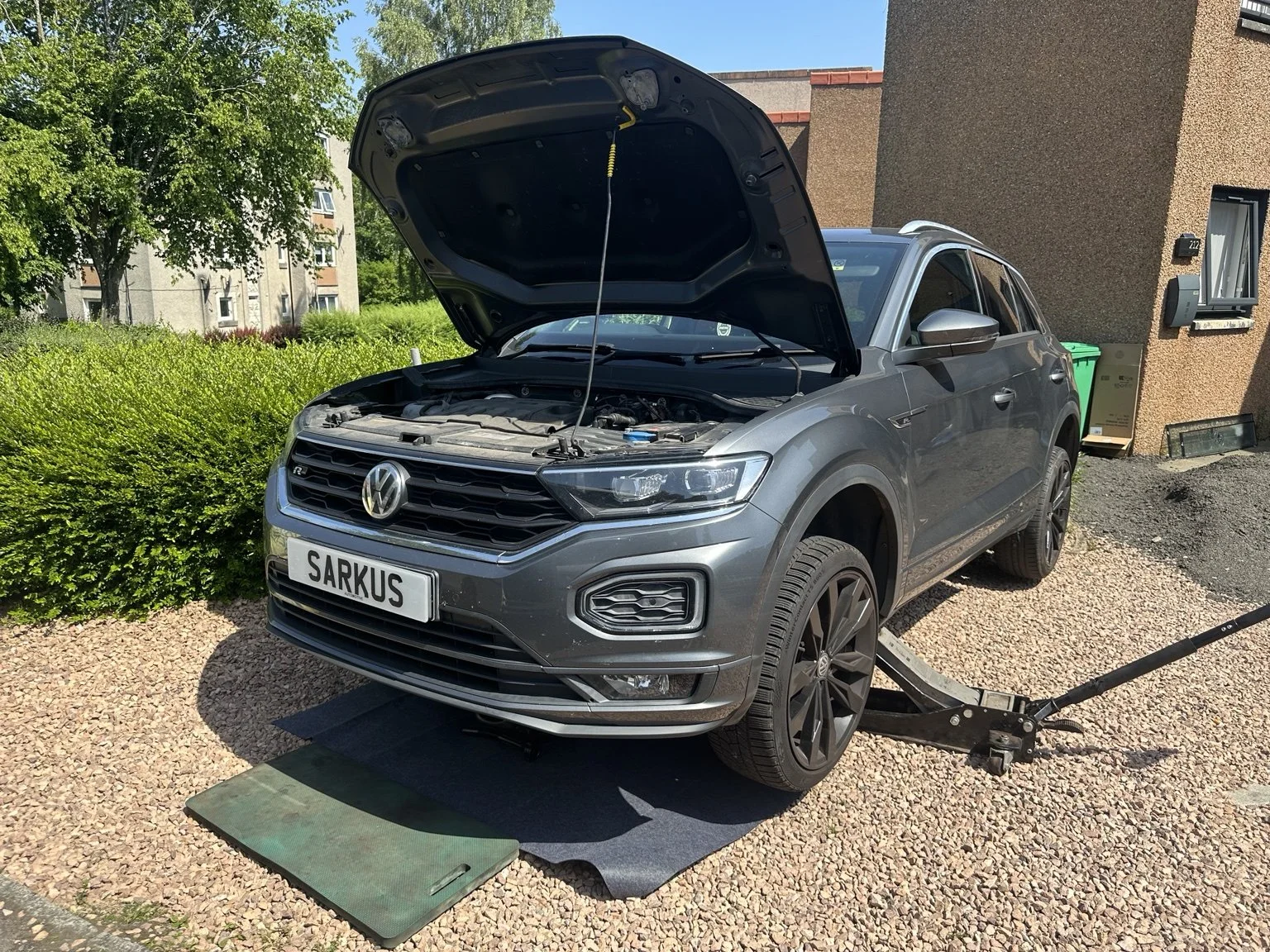 A gray Volkswagen SUV with an open hood, positioned on a car lift in an outdoor setting, indicating maintenance or repair work.
