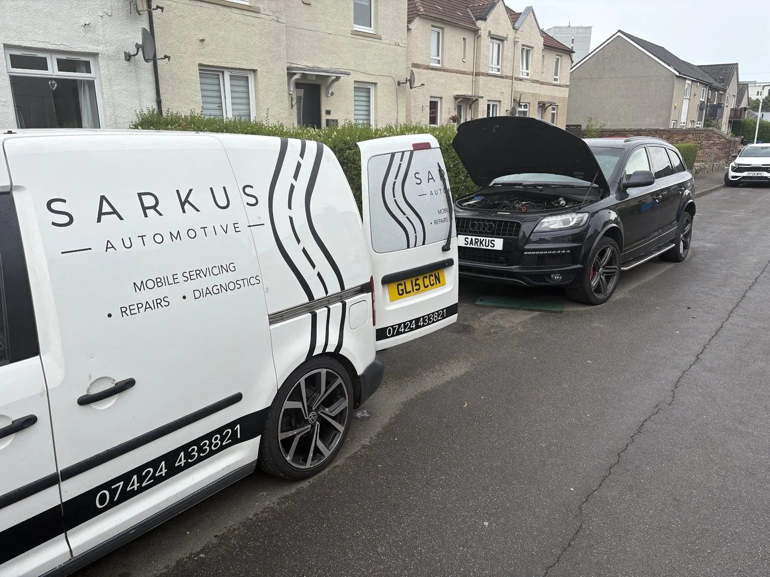 A black Audi SUV with its hood open parked on a residential street next to a white mobile servicing van labeled "Sarkus Automotive" which offers repairs, diagnostics, and mobile servicing. The van has a UK license plate and phone number.
