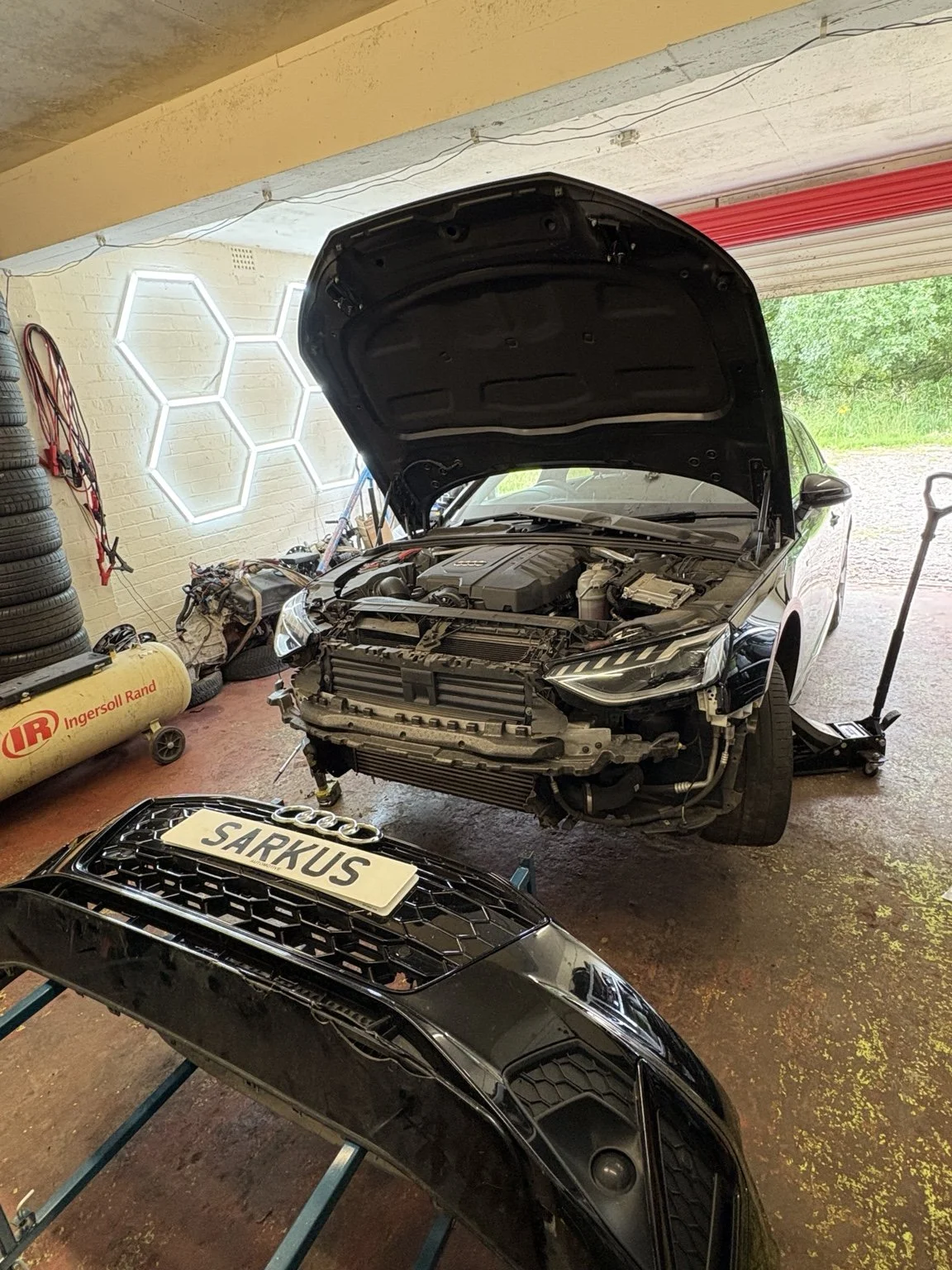 A black Audi car with a custom license plate 'SAKUS' is on a lift in a garage, with its front bumper removed and the hood open, exposing the engine. The garage has a wall with hexagonal light fixtures, stacked tires, and a yellow air compressor.