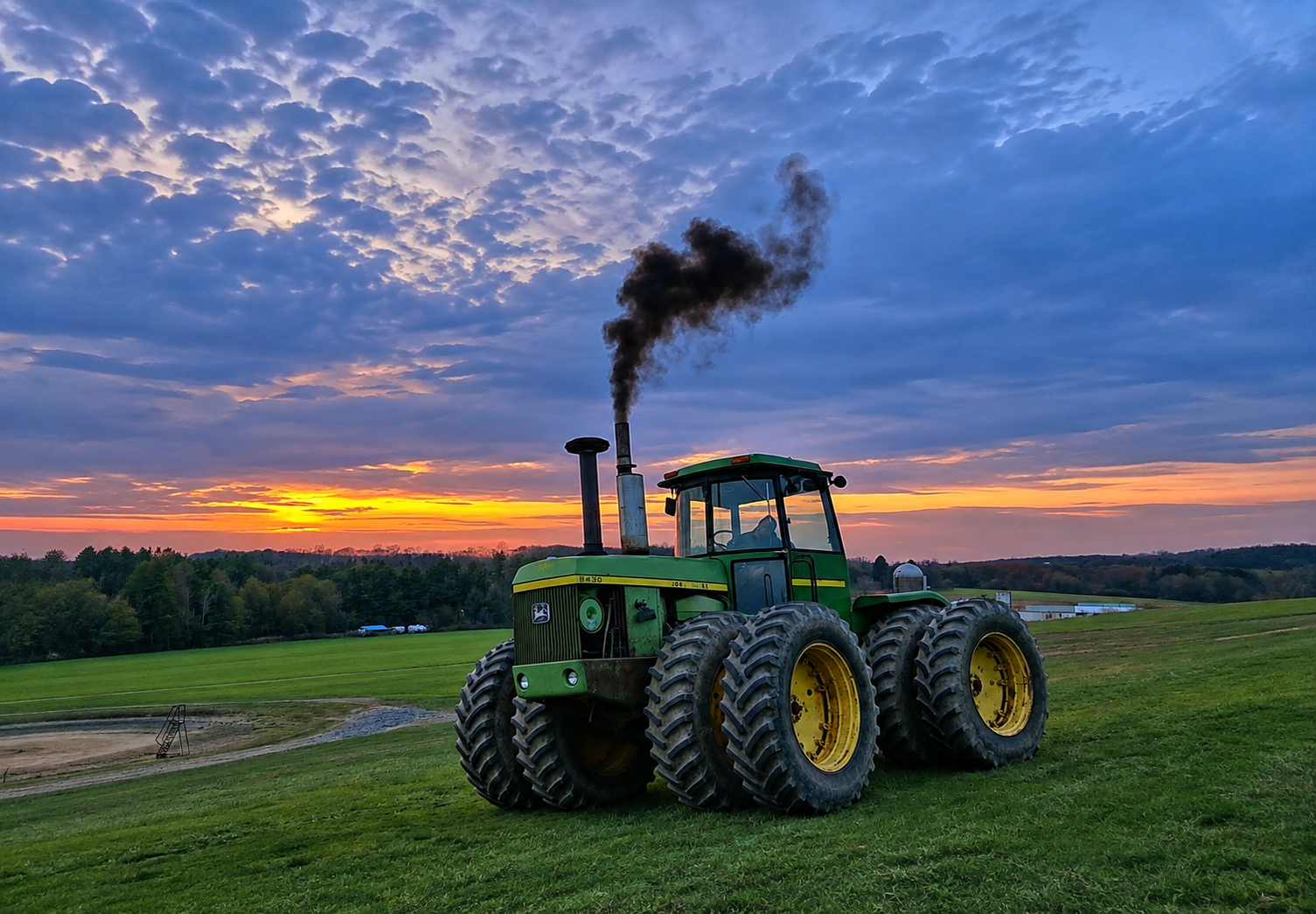 Green tractor on a grassy field at sunset with clouds and smoke coming from its exhaust pipe.