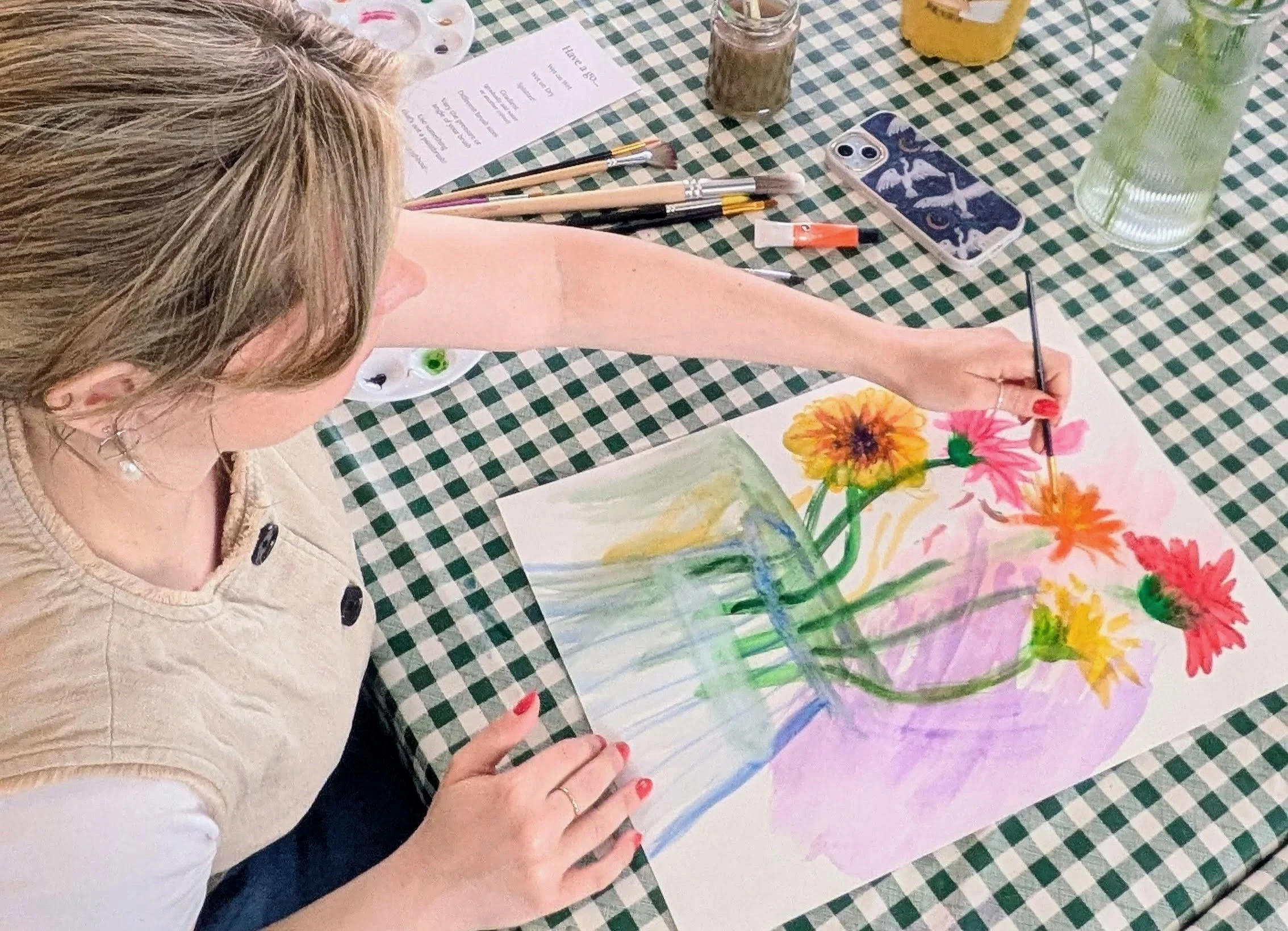 A person with blonde hair painting colorful flowers on paper at a table with a green and white checkered tablecloth.