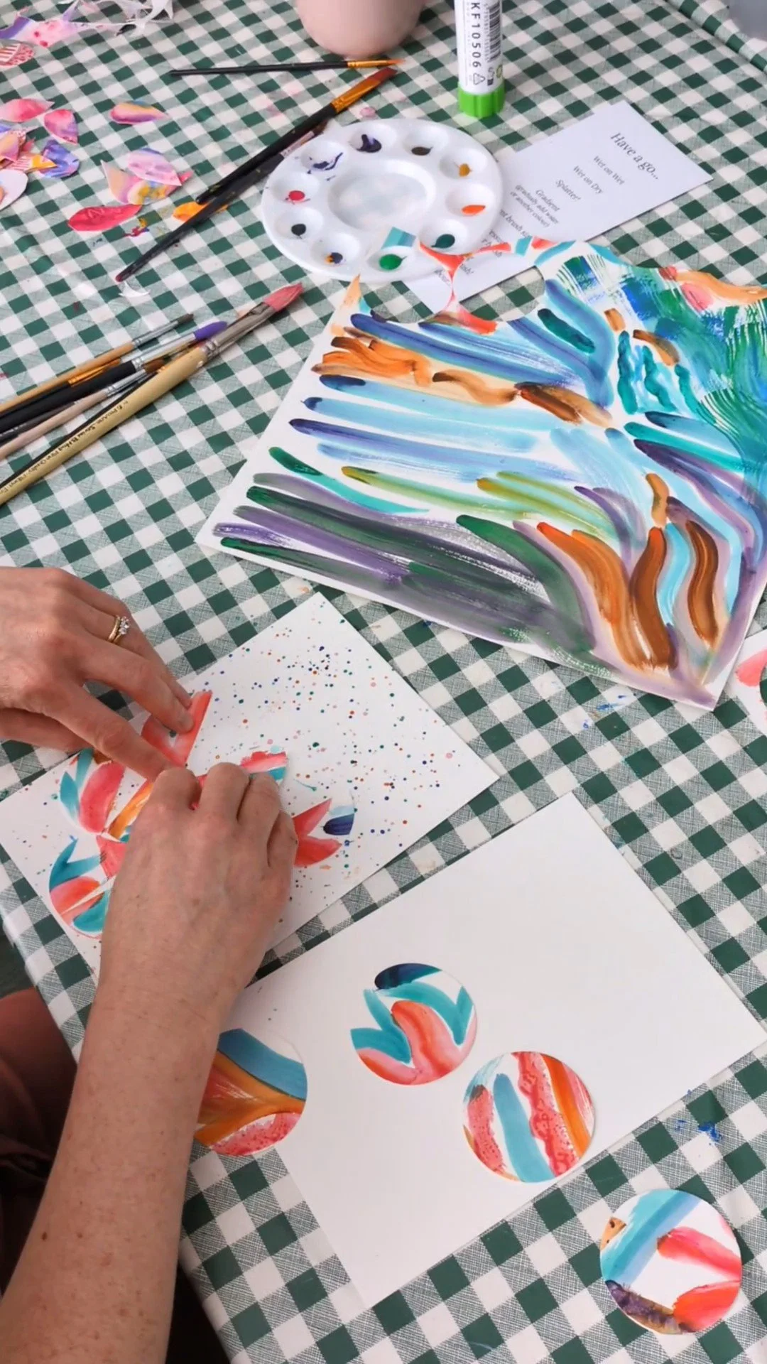 A person is creating colorful abstract art of a landscape in greens, blues and oranges. She is making a collage from cut-out circular shapes using watercolor paints and brushes at a craft table.