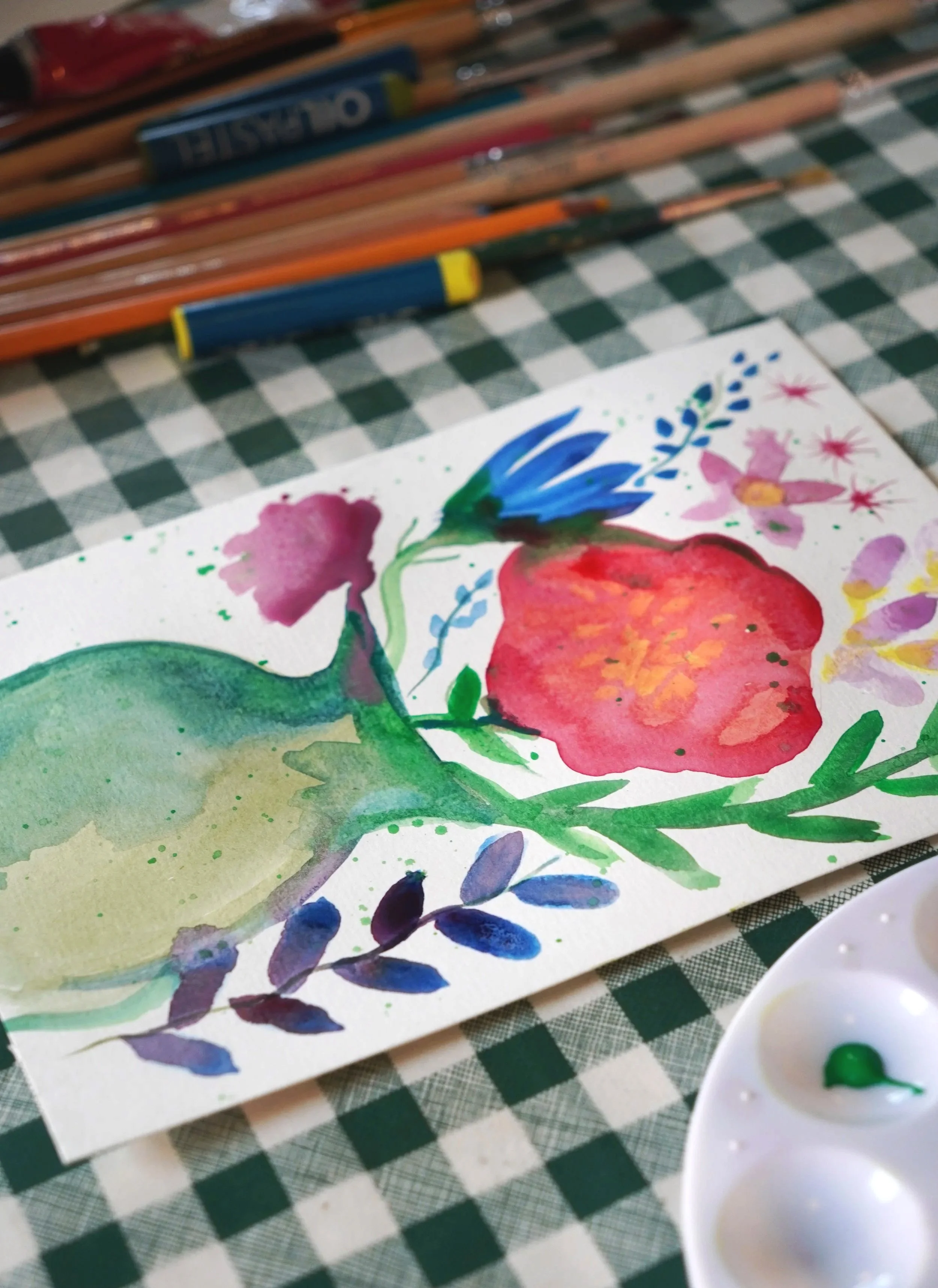 Close-up of a watercolor painting featuring vibrant floral designs and leaves on a piece of textured paper, with a palette of green and blue paints nearby, placed on a checkered tablecloth.