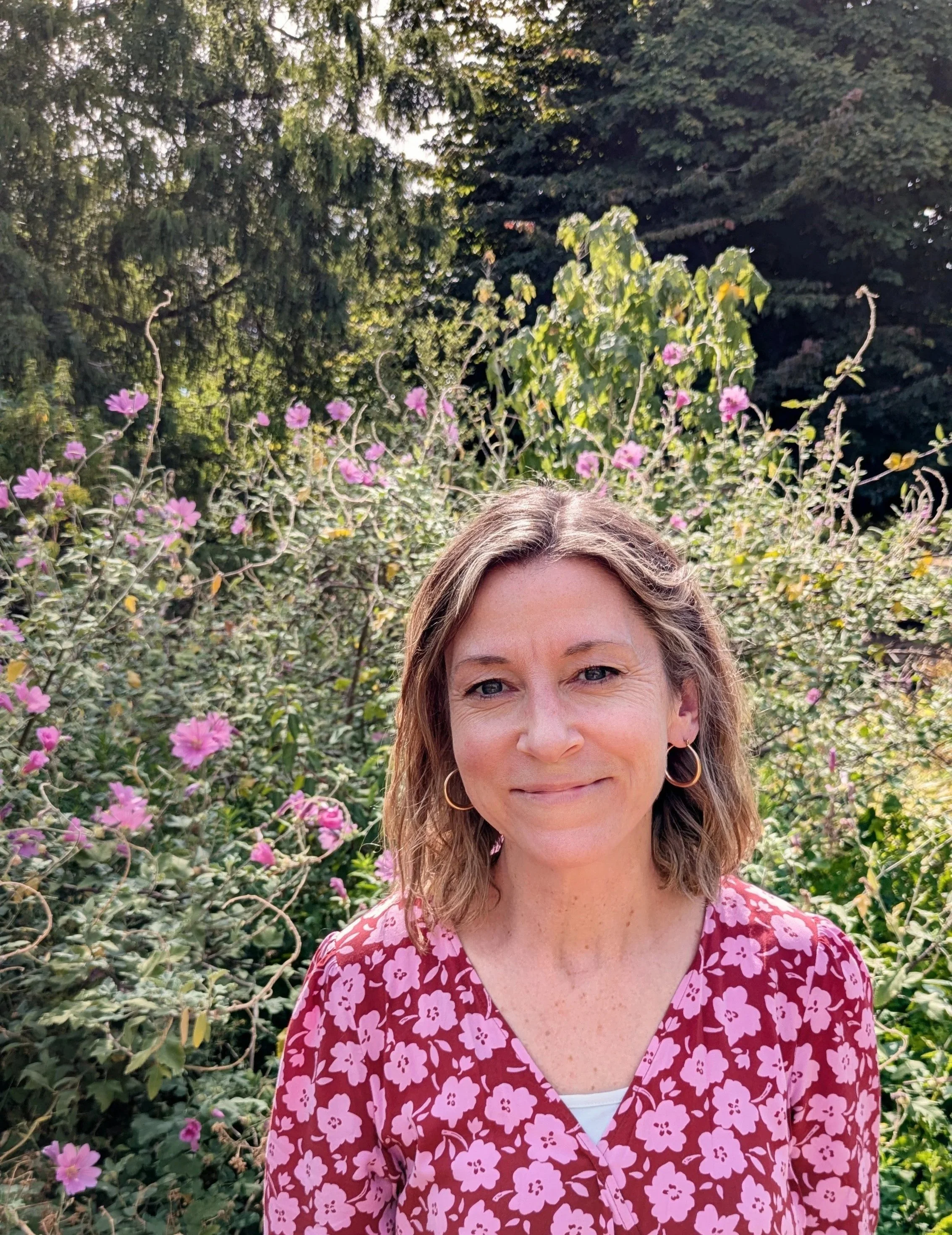 Close-up of Laura Dampney, a smiling woman with shoulder-length brown hair wearing gold hoop earrings and a red dress with pink and white floral pattern, standing in front of a garden with pink flowers and green leafy trees in the background.