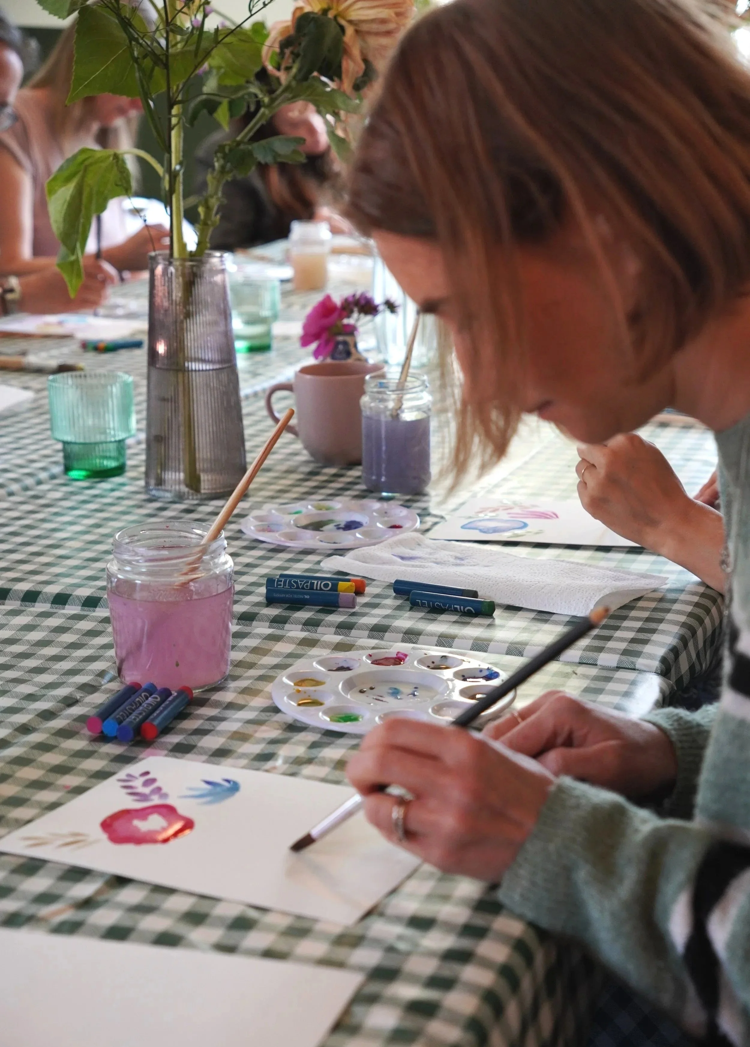 People sitting around a table engaging in watercolor painting of flowers, with paint palettes, brushes, and art supplies visible on a checkered tablecloth.