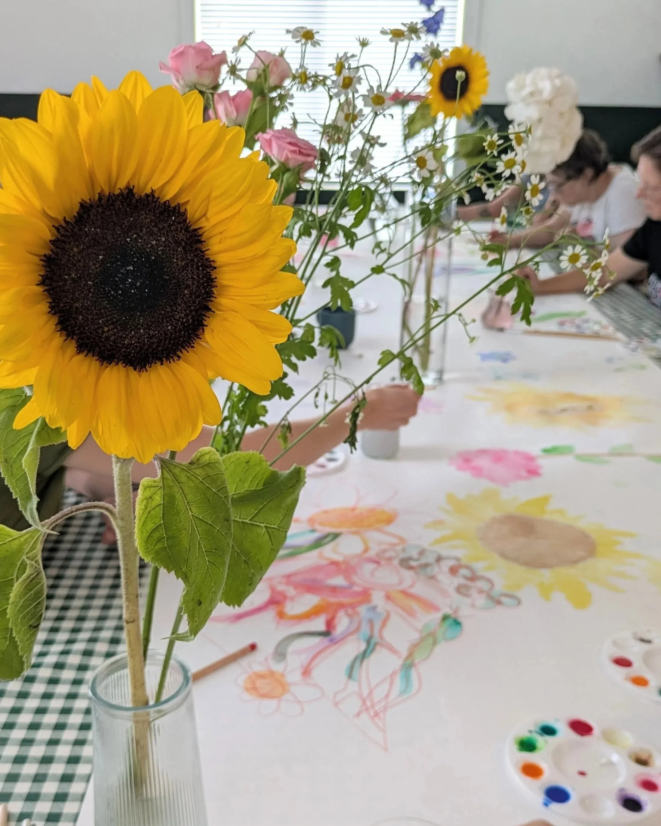 A close-up flower arrangement with a large sunflower, roses, daisies and blue flowers in a glass vase on a table covered with a large sheet of paper.  Two people in the background are engaged in painting large flowers with expressive brushstrokes.