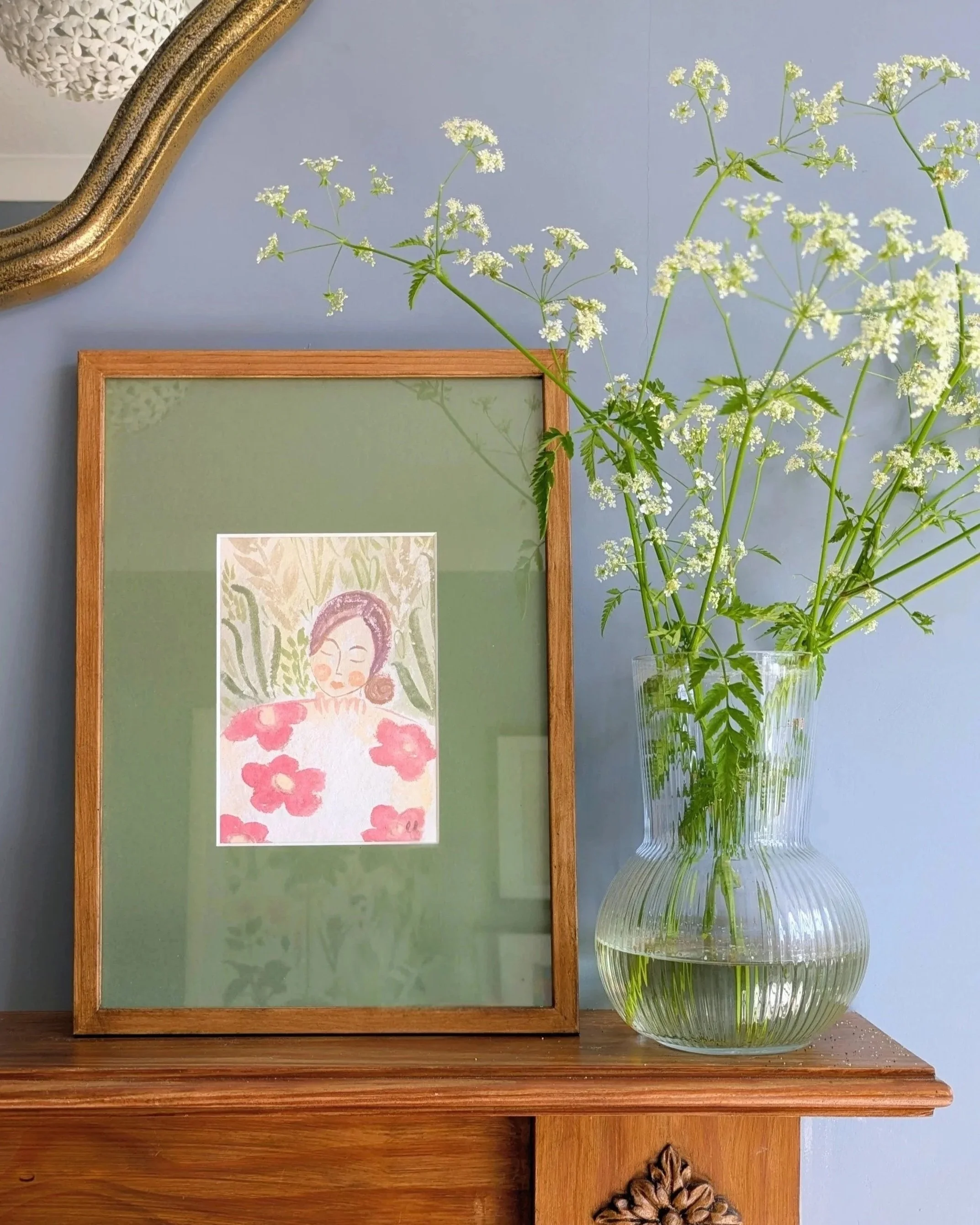A small painting of a peaceful woman wearing red flowers in a garden surrounded by green leaves. Set in a wooden frame with a wide green mount. The painting is on a wooden fireplace next to a glass vase containing cow parsley.